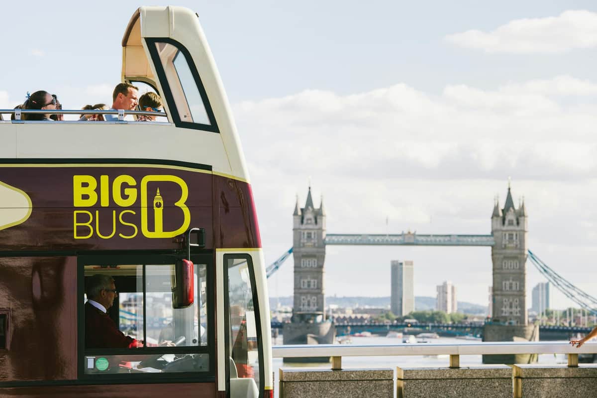 Bus touristique à deux étages avec le logo "BIG BUS", personnes sur le pont supérieur, Tower Bridge en arrière-plan sous un ciel partiellement nuageux.