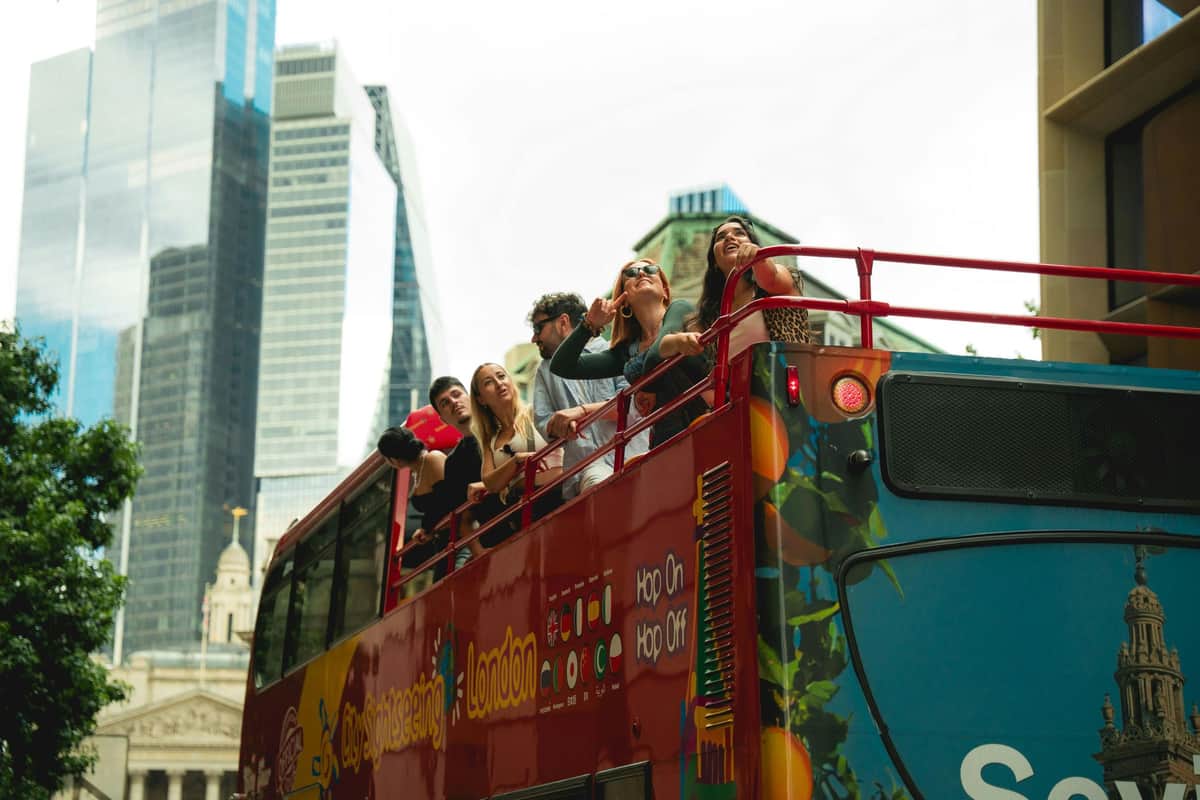 Un groupe de personnes sur le pont supérieur d'un bus touristique rouge dans une ville avec des gratte-ciel modernes et une architecture plus ancienne.