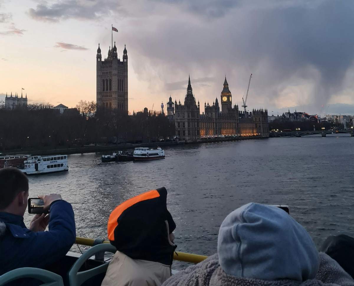 Londres : Visite nocturne en bus Des personnes chaudement vêtues sur un bateau, observant Big Ben et le Palais de Westminster illuminés sur la Tamise au crépuscule.