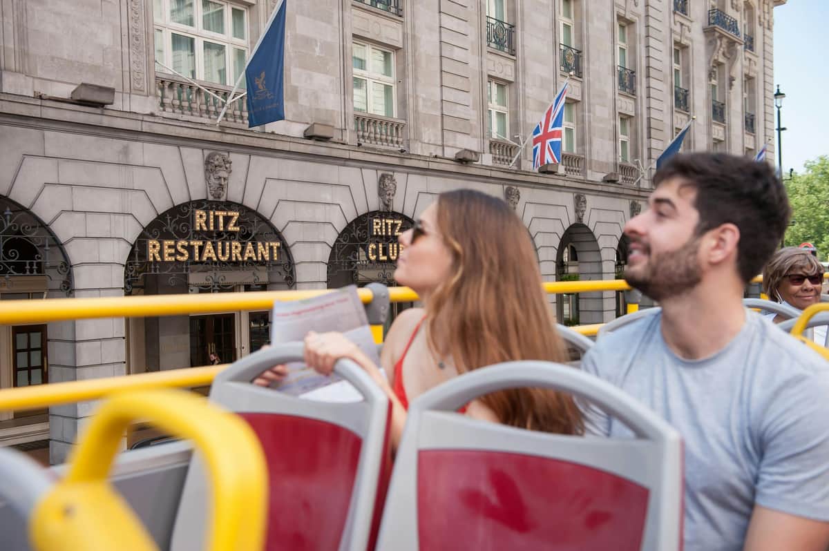 Deux personnes assises dans un bus touristique à impériale, avec le bâtiment du Ritz Club et le drapeau Union Jack visibles à l'arrière-plan.