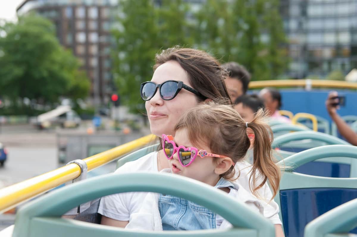 Tootbus Londres : Visite pour enfants Une femme portant des lunettes de soleil foncées et un enfant portant des lunettes de soleil roses en forme de cœur montent dans un bus à toit ouvert, avec des arbres et des bâtiments flous en arrière-plan.