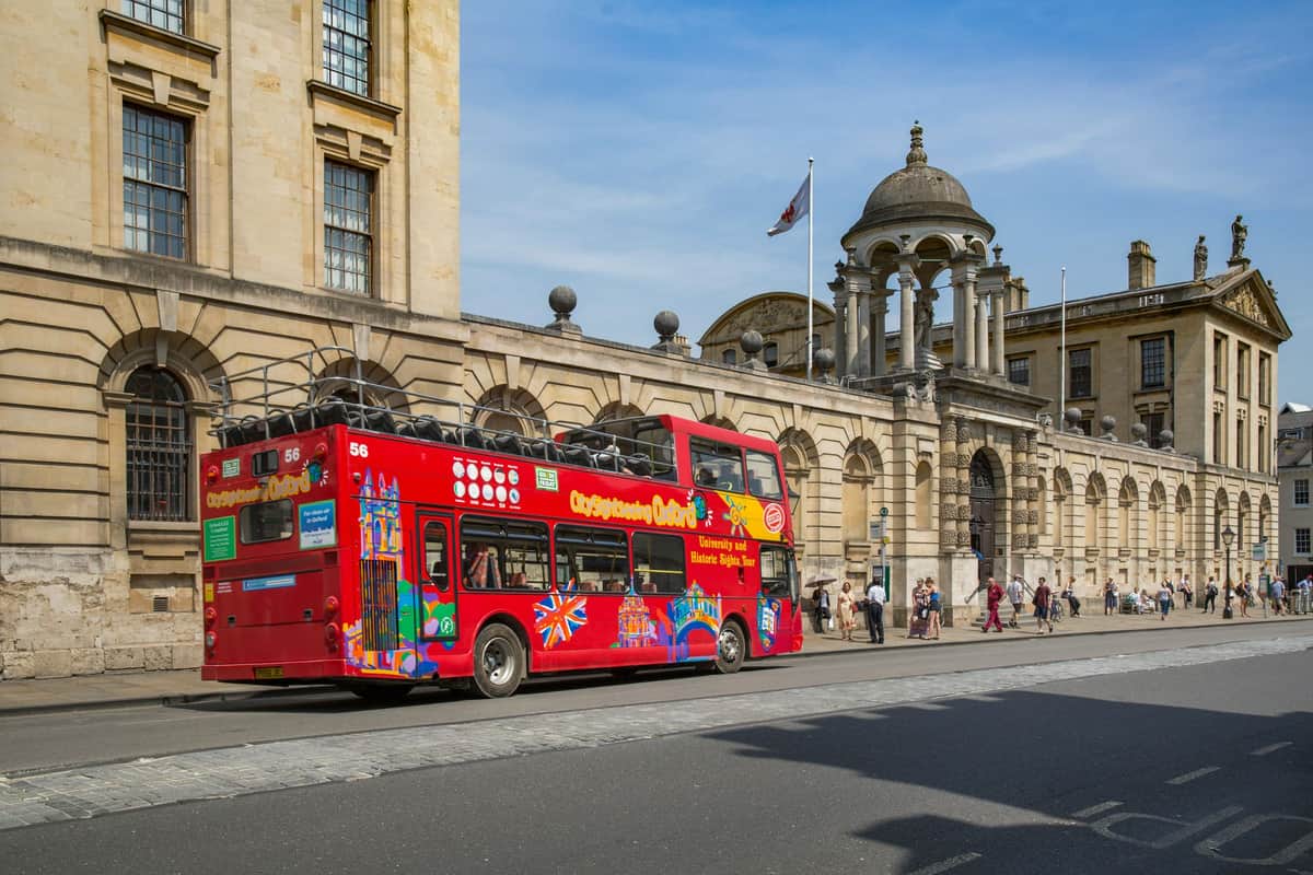 Un bus touristique rouge à impériale passe devant un bâtiment historique en pierre avec des fenêtres arquées et une structure en forme de dôme. Des passants se trouvent à proximité.