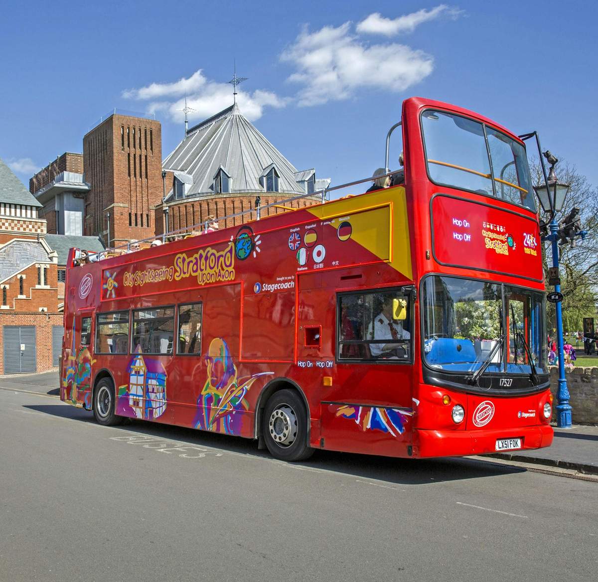 Un bus touristique à impériale, rouge vif, portant l'inscription "City Sightseeing Stratford", stationné dans une rue à proximité de bâtiments architecturaux.