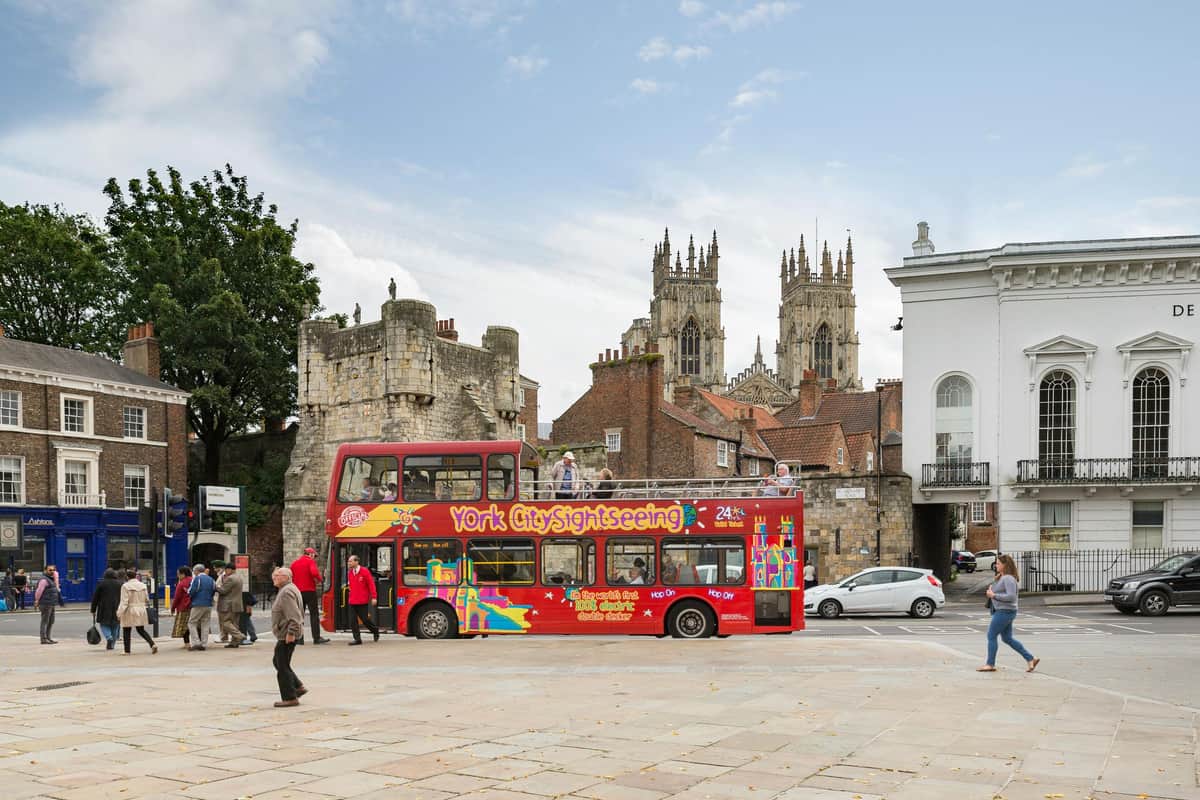 Bus hop-on hop-off York Un bus touristique à impériale portant l'inscription "York City Sightseeing" à proximité de murs médiévaux et de personnages, avec York Minster en arrière-plan.