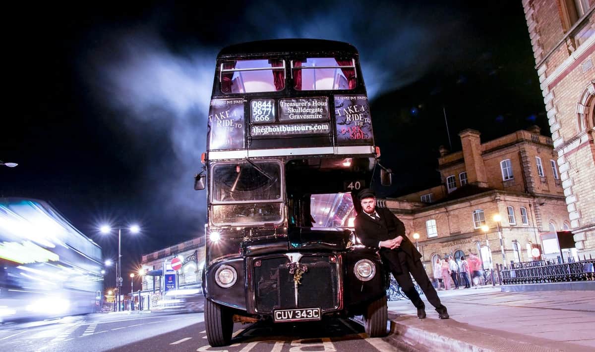 Un homme en costume noir s'appuie sur l'avant d'un bus noir à impériale, la nuit, avec des bâtiments urbains illuminés en arrière-plan.