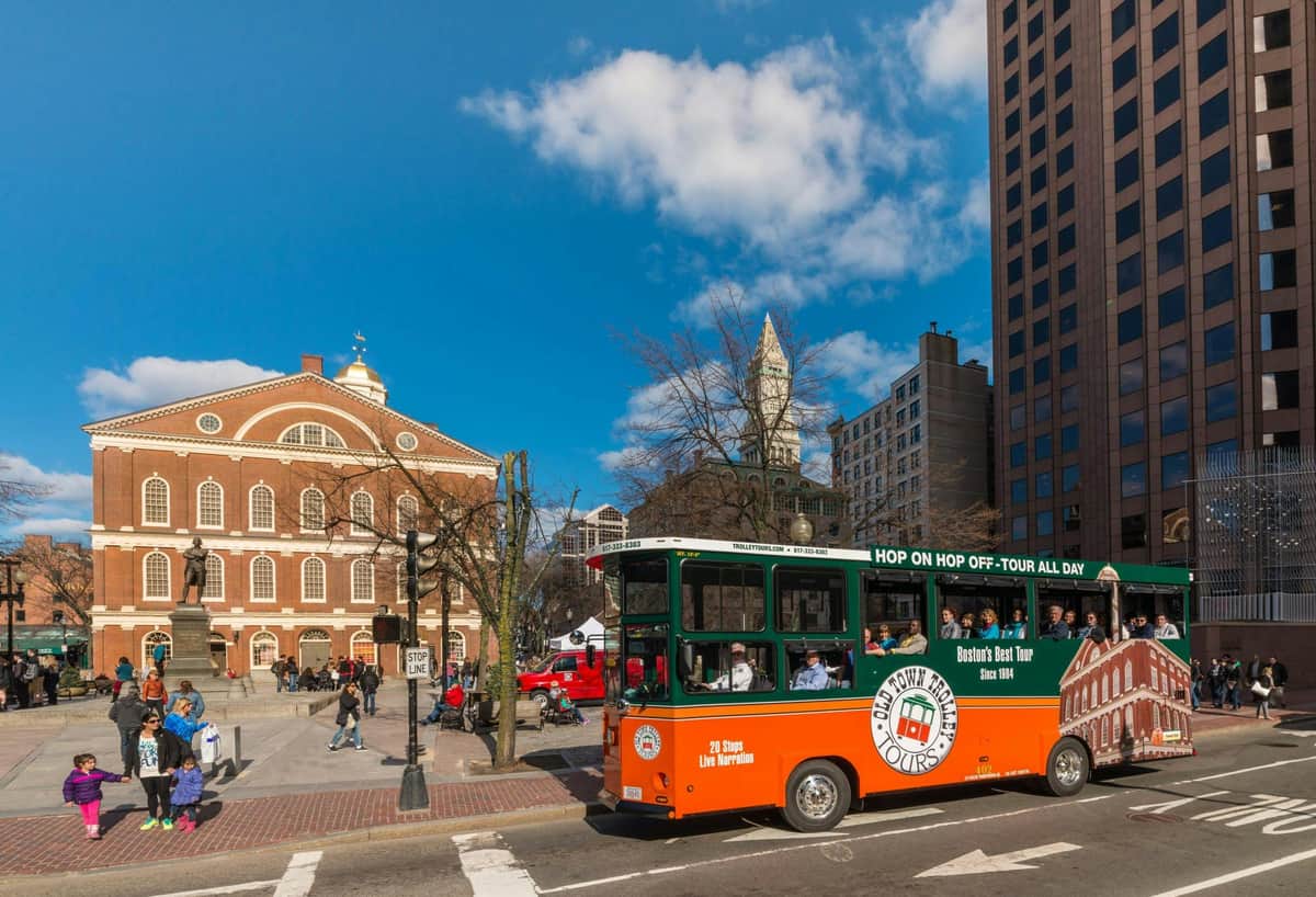 Un bus touristique orange et vert traverse une place de la ville avec des bâtiments historiques en briques et une tour d'horloge sous un ciel bleu.