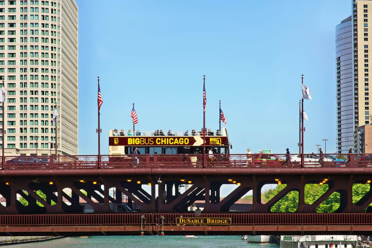 Un bus touristique à impériale portant l'inscription "BigBus Chicago" traverse le pont DuSable, orné de drapeaux américains, sous un ciel bleu limpide.