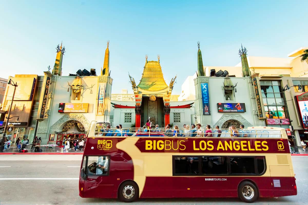 Bus de tourisme à deux étages devant le Grauman's Chinese Theatre à Los Angeles, avec des personnes sur le pont supérieur et un ciel bleu clair.