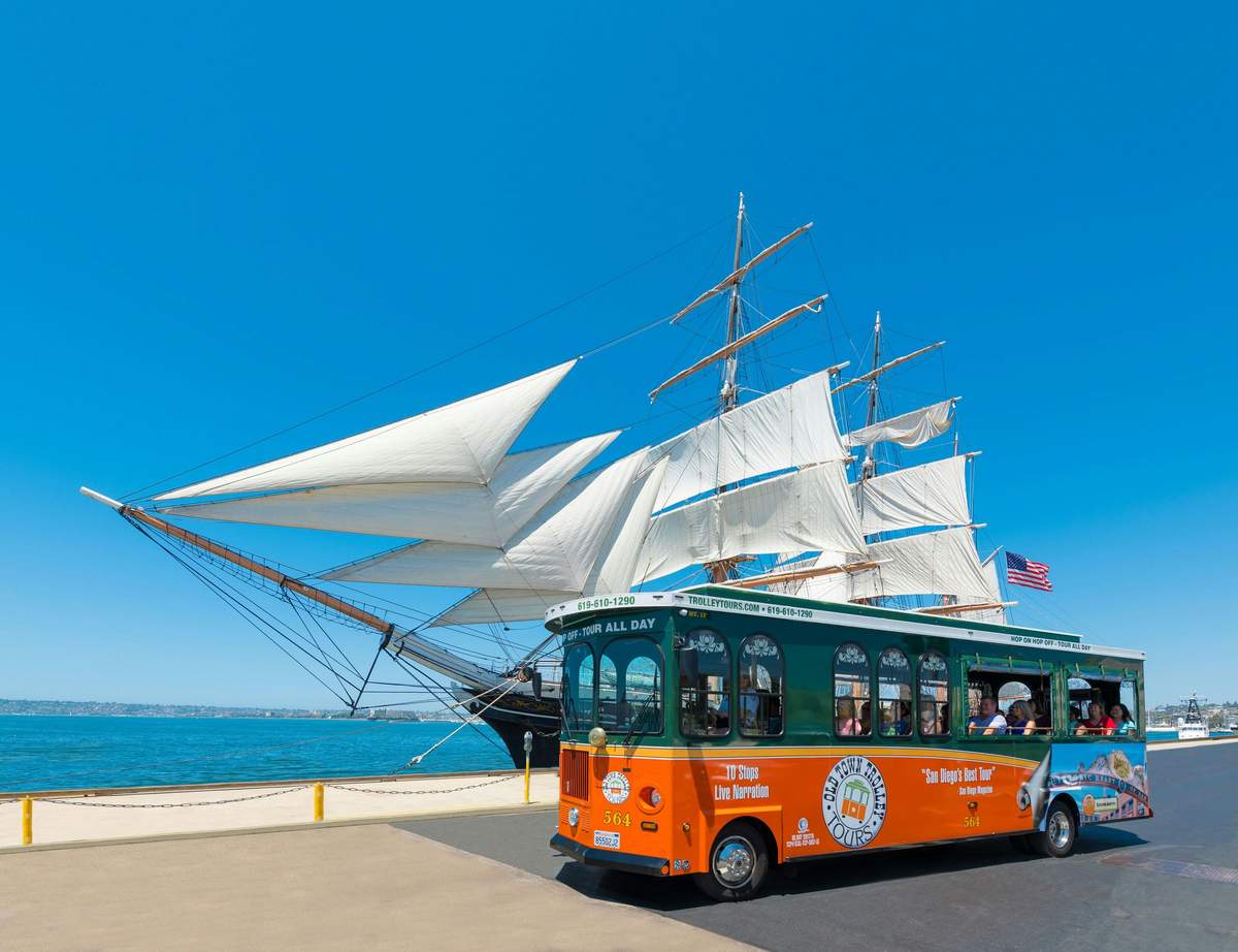 Un trolley vert et orange passe devant un grand voilier aux voiles blanches près d'un front de mer sous un ciel bleu clair.