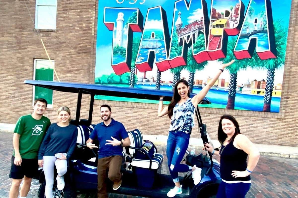 Trois personnes posent joyeusement devant une grande peinture murale colorée "Tampa". Elles se tiennent à côté d'une voiturette de golf.