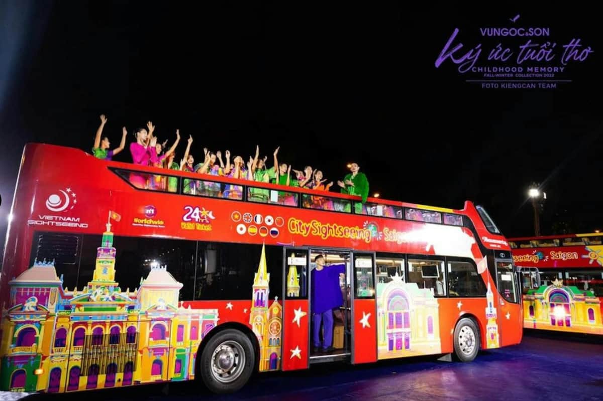 Un bus touristique à deux étages aux couleurs vives, avec des passagers sur le pont supérieur ouvert qui font des signes excités la nuit.