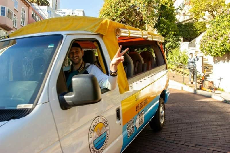 San Francisco: Guided Open-Air Bus Tour (San Francisco) A man in a white van with yellow accents, smiling and making a peace sign, parked in a sunny urban area.