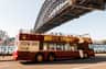 A red double-decker tour bus labeled "Big Bus Sydney" under Sydney Harbour Bridge, with seated passengers on the upper deck.
