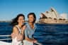 Two women smiling on a boat with water and the Sydney Opera House in the background under a clear blue sky.