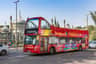 Red double-decker sightseeing bus in Brighton with tourists on the top deck, passing by a building with domed architecture.