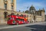 A red double-decker sightseeing bus drives by a historic stone building with arched windows and a domed structure. Passersby nearby.